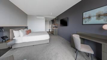 A modern hotel room with a bed, bedside table, desk, chair, TV, and artwork on the wall.