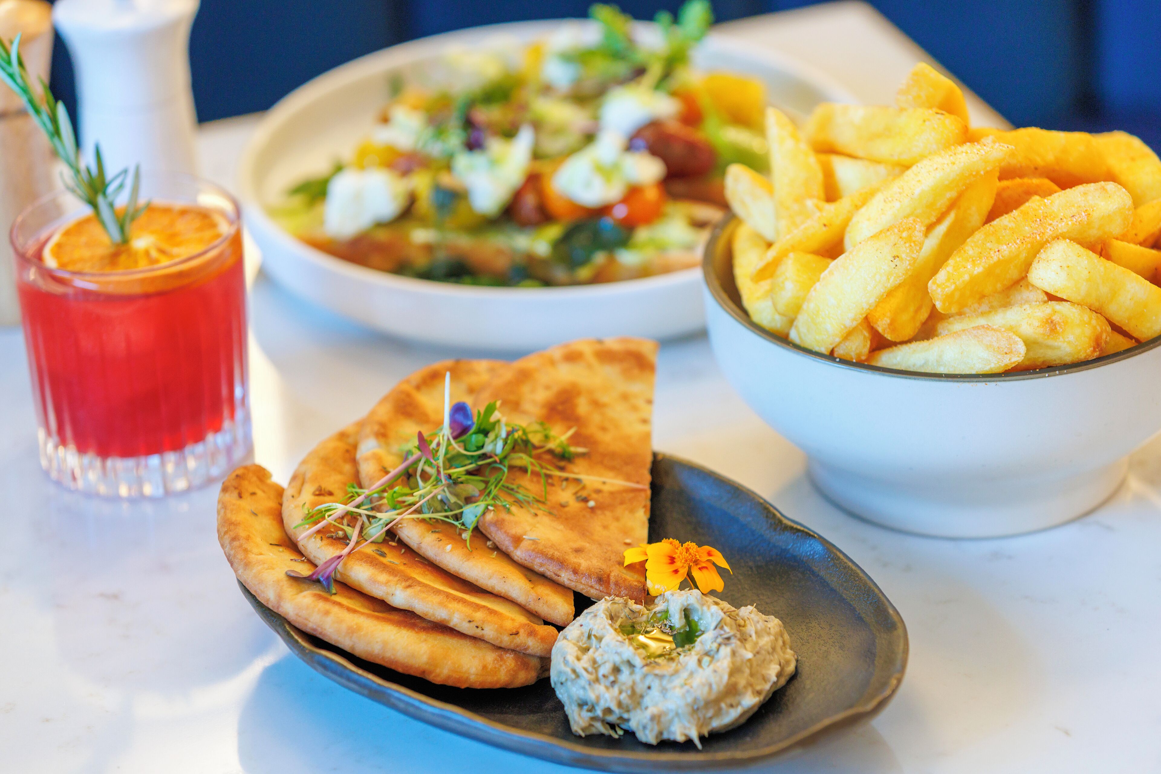 A table with a plate of flatbread, a bowl of fries, a glass of red drink, and a bowl of salad.