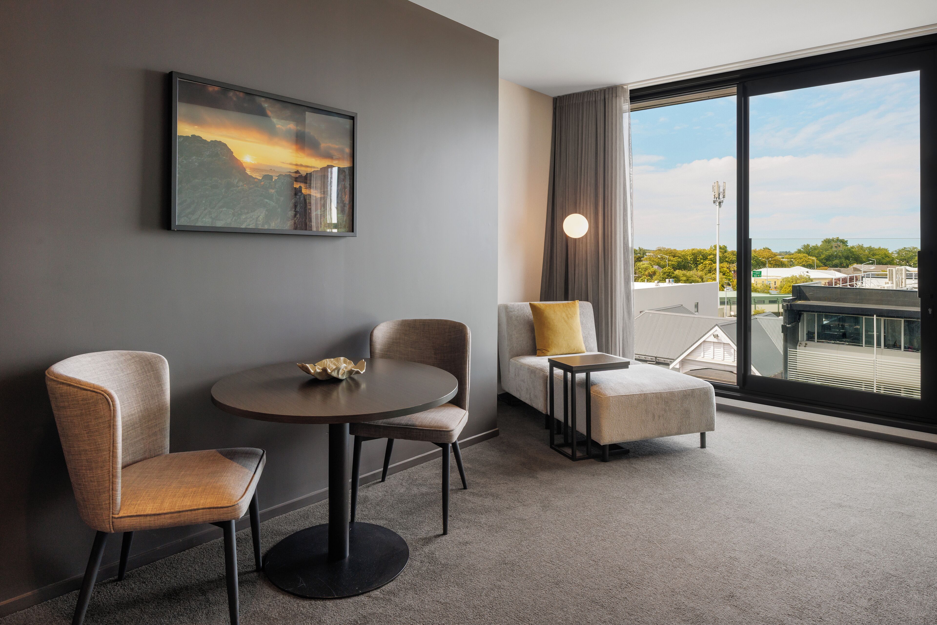 Une chambre d’hôtel moderne dotée d’une grande fenêtre, d’une table ronde, de deux fauteuils et d’un canapé agrémenté d’un coussin jaune.