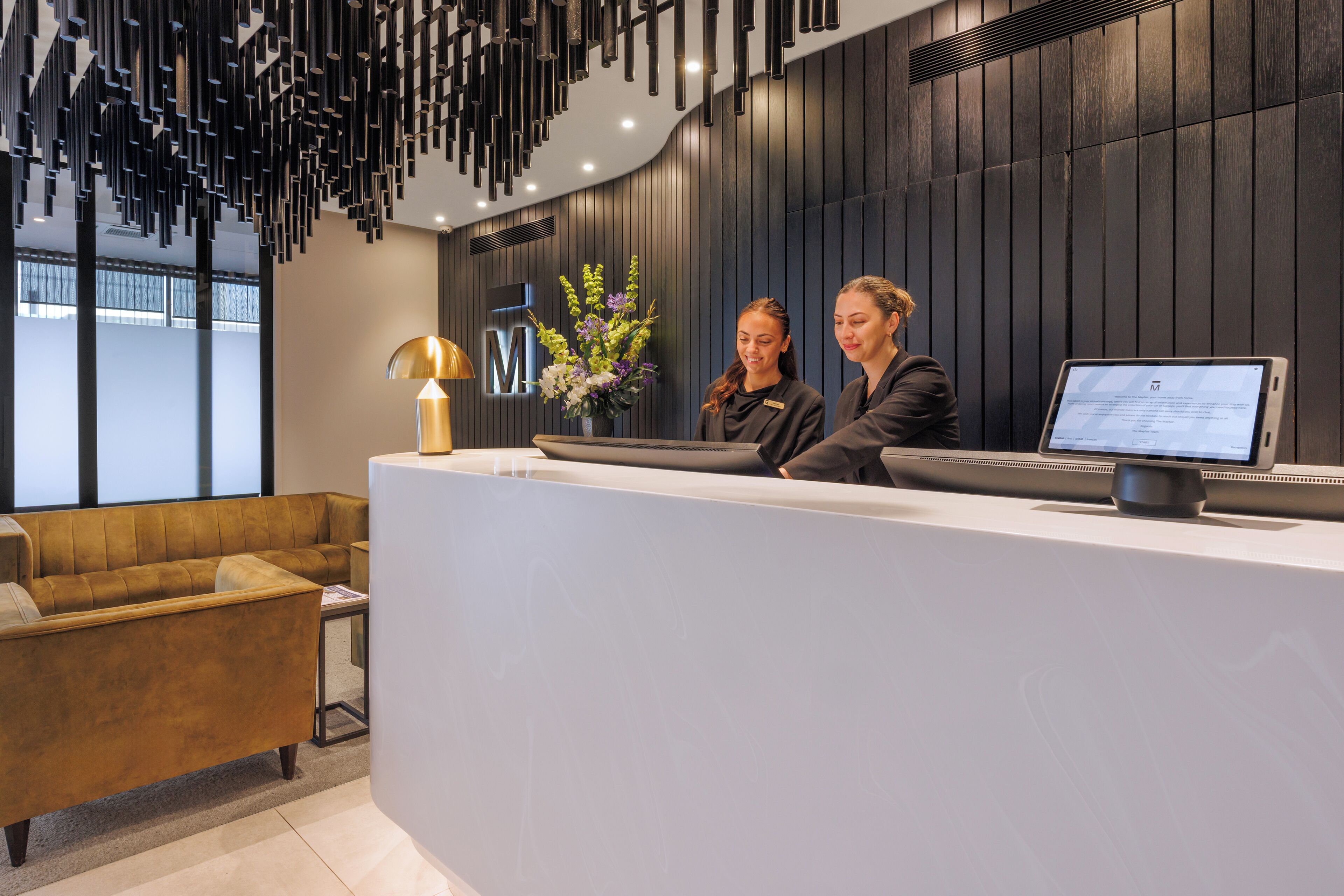 Un comptoir d’accueil avec deux employés, une lampe moderne et un arrangement floral.