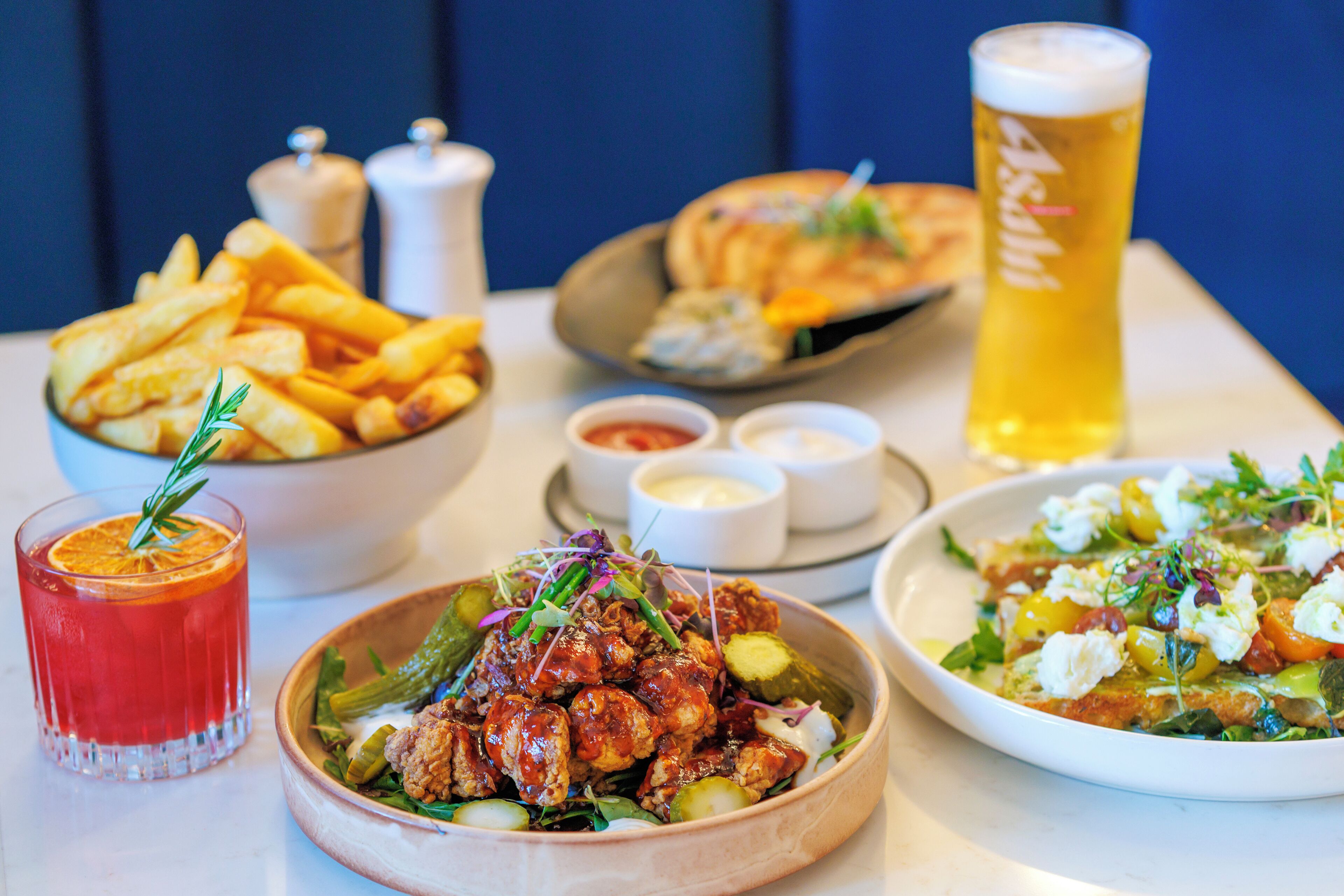 Una mesa con varios platos, incluyendo papas fritas, una ensalada y un recipiente con comida, además de una cerveza y una bebida.