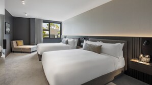 A modern hotel room with two large beds, a sofa, a bedside table with a phone, and a window with a view of the outside. - The Mayfair (Christchurch)