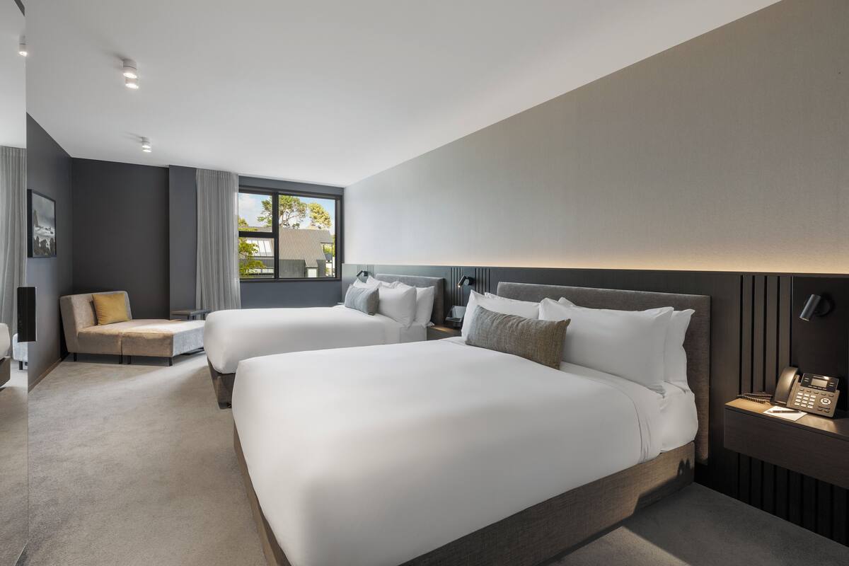 A modern hotel room with two large beds, a sofa, a bedside table with a phone, and a window with a view of the outside.