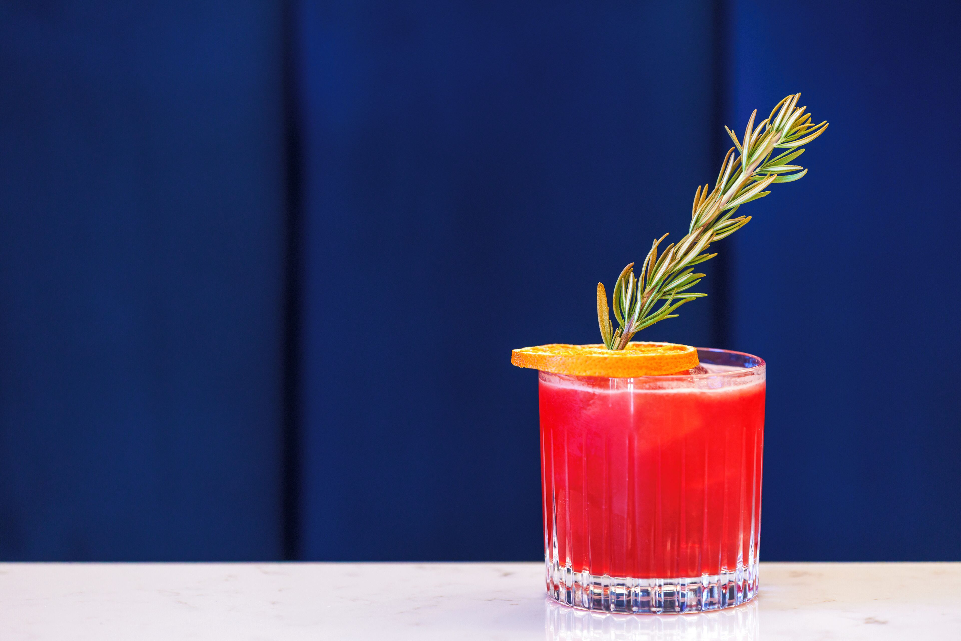Un verre d’une boisson rouge, agrémenté d’une tranche d’orange et d’une branche de romarin, posé sur une surface blanche, le tout sur un fond bleu.