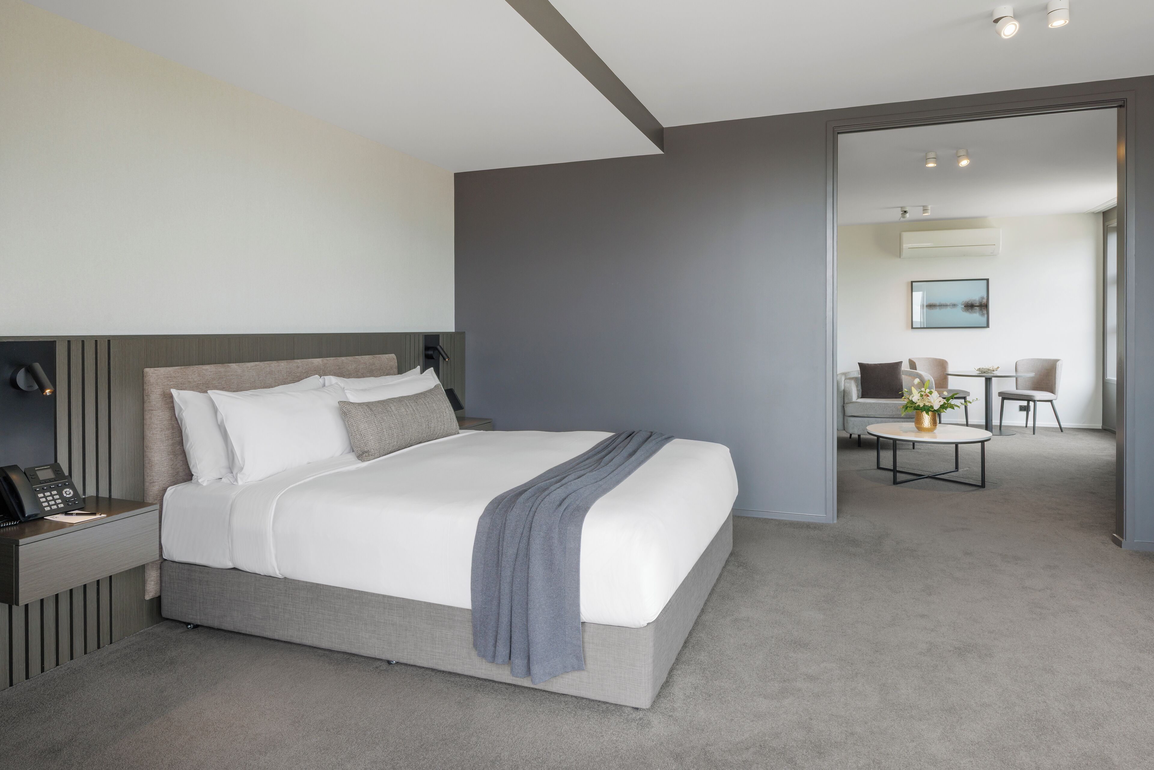 A modern hotel room with a large bed, bedside table with a phone, and a view into a dining area.