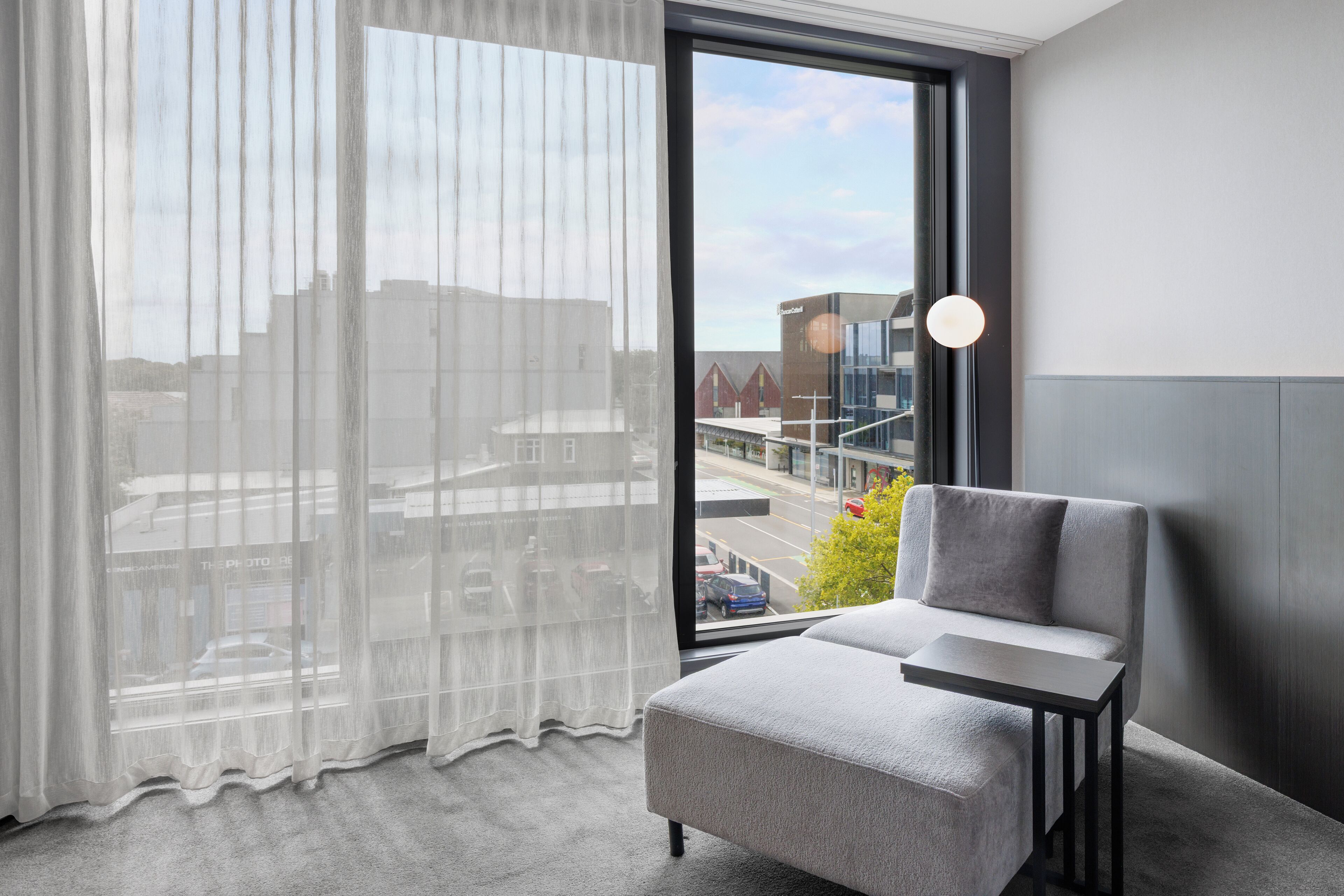 A modern hotel room with a large window, a grey armchair, and a small table.