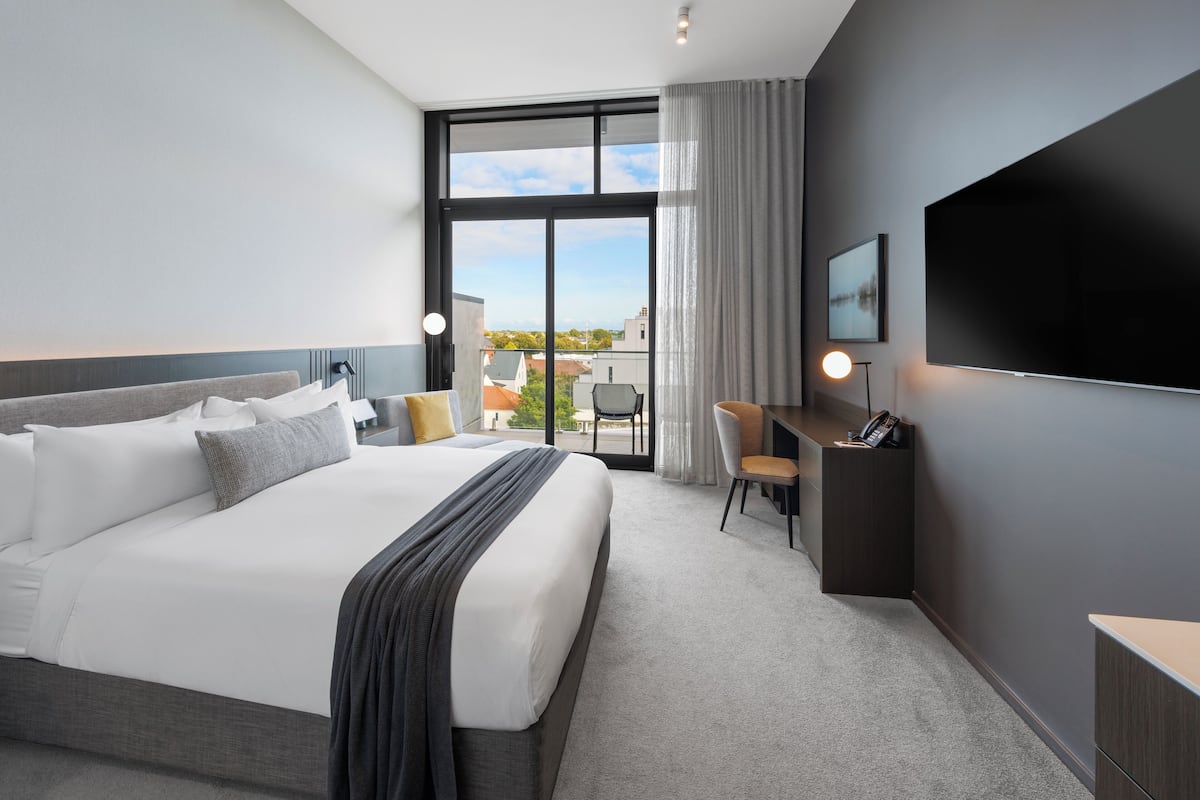 A modern hotel room with a large bed, a desk, a chair, a TV, and a view of the city.