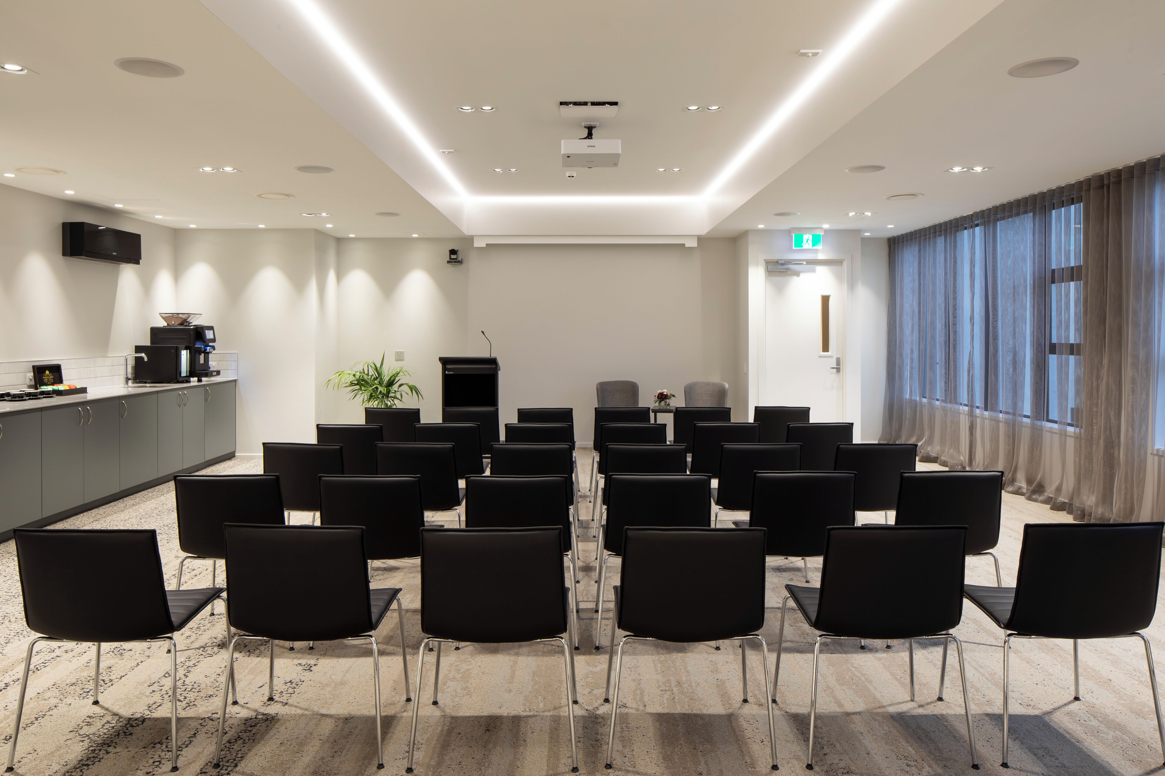 A conference room with rows of black chairs, a kitchen area, and a large window with curtains.