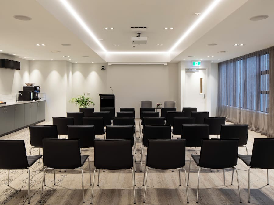 A conference room with rows of black chairs, a kitchen area, and a large window with curtains.