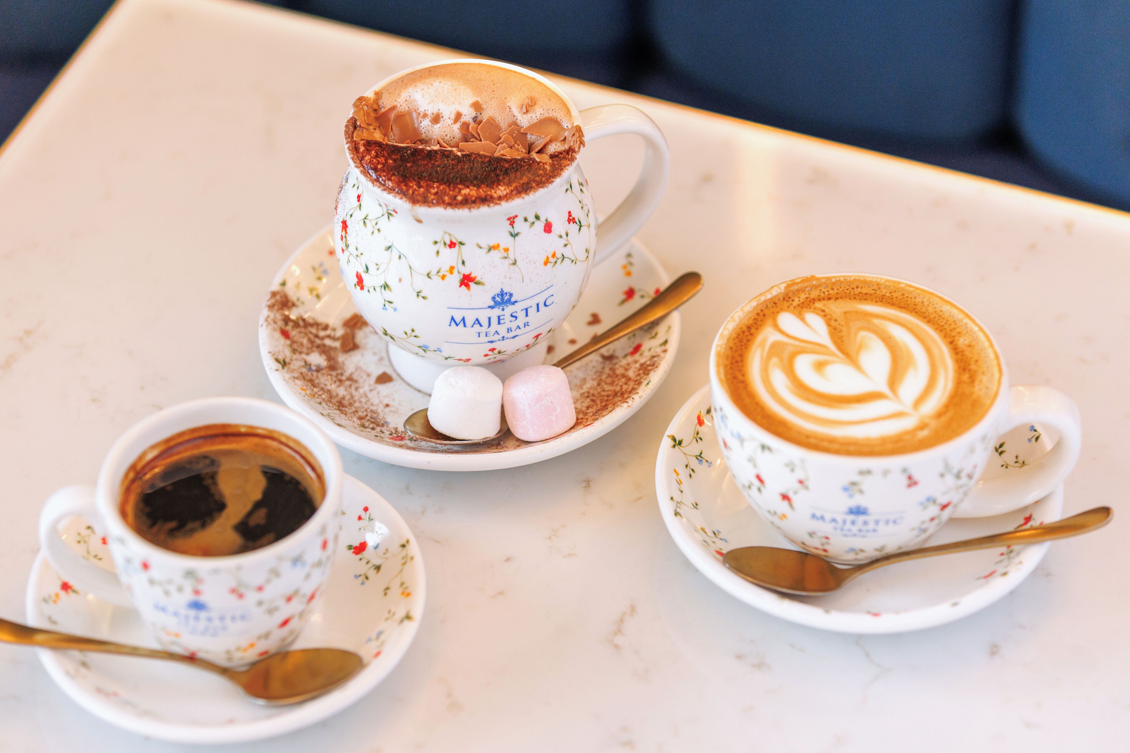 A table with three cups of coffee, each with a floral pattern and the name "MAJESTIC TEA BAR" on the cup. There are marshmallows and a spoon on the saucer of the middle cup.