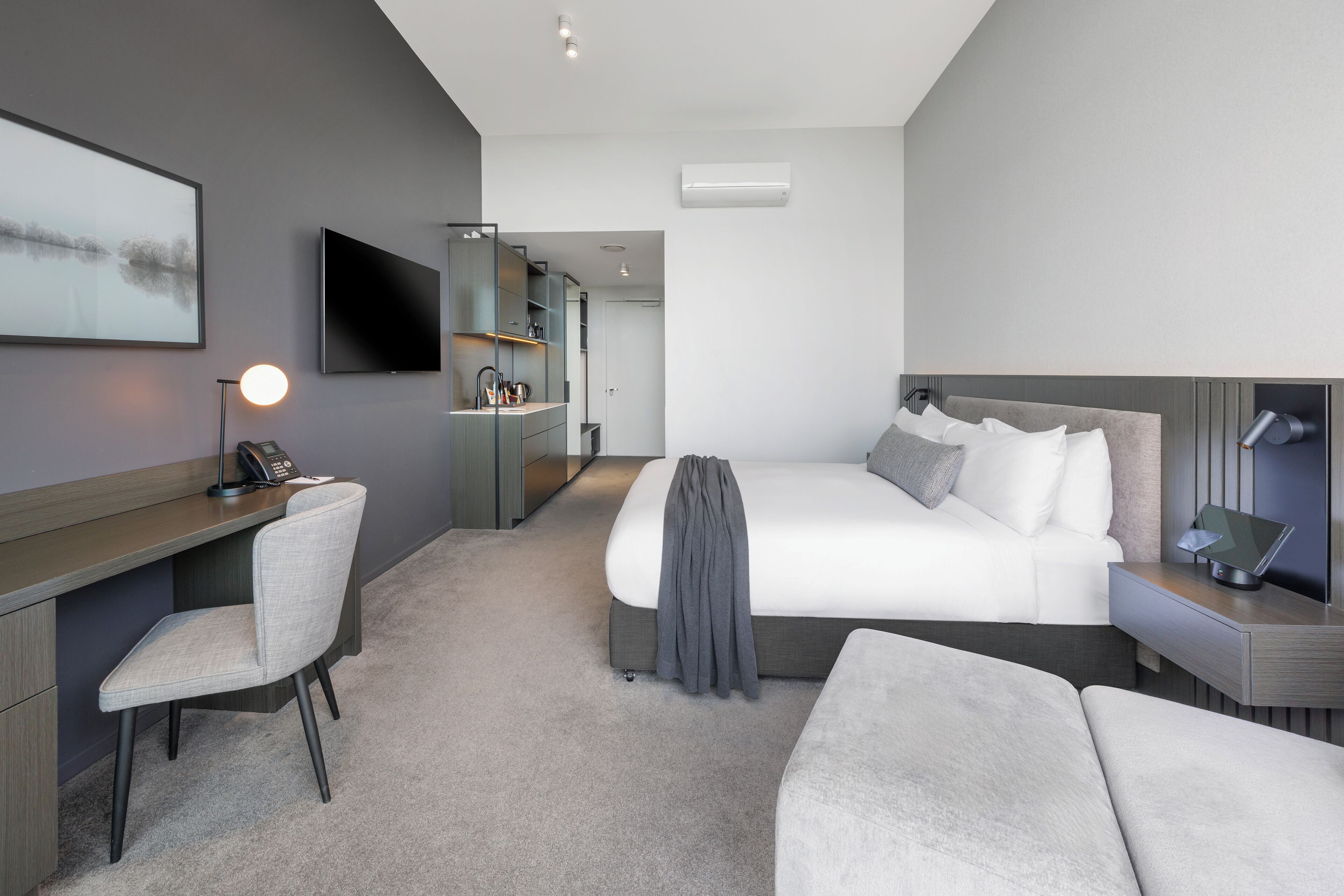 Una habitación de hotel moderna con cama, escritorio, silla, televisor y una kitchenette pequeña.