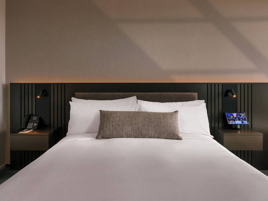Een keurig opgemaakt bed met witte lakens, een bruin kussen en een nachtkastje met een telefoon en een digitale klok.