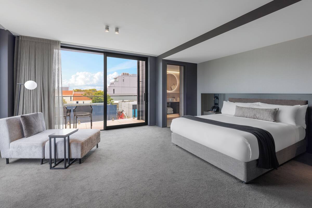 A modern hotel room with a large bed, a sofa, a small table, and a view of the city through large windows.