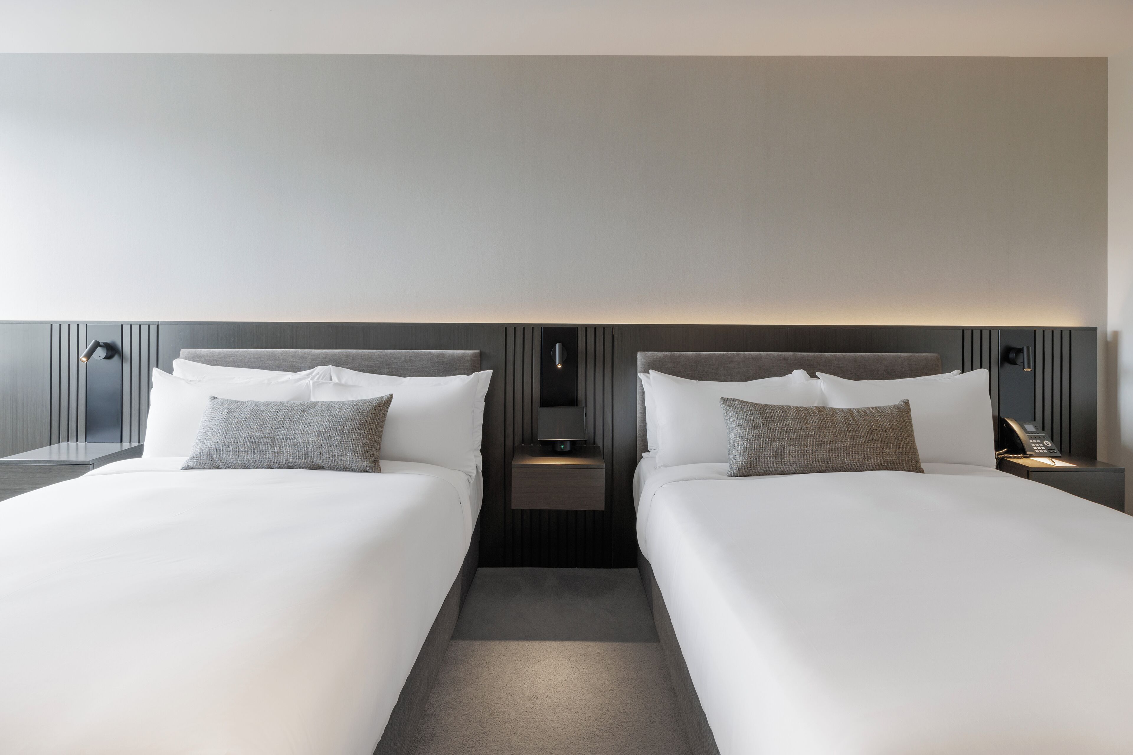 A modern hotel room with two beds, a bedside table with a lamp, and a small nightstand with a phone.