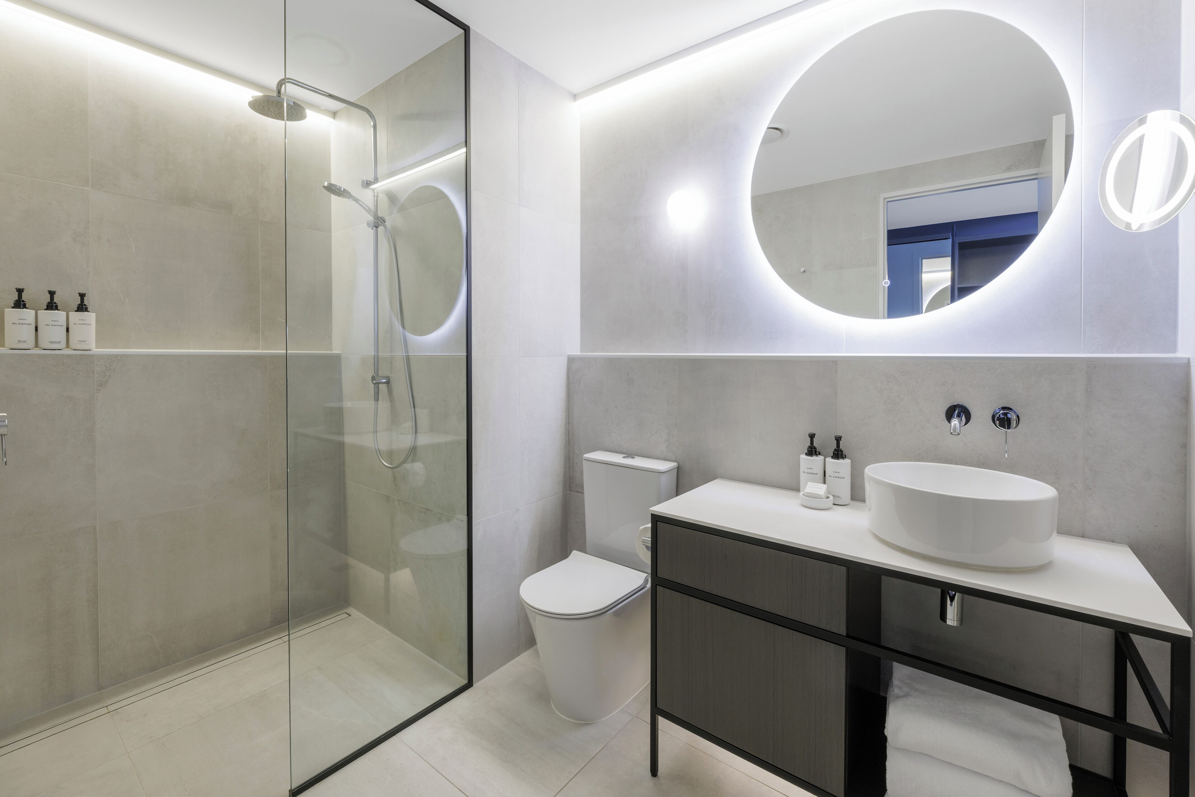 Un baño moderno con cabina de ducha de vidrio, inodoro blanco y tocador con espejo redondo y lavamanos.
