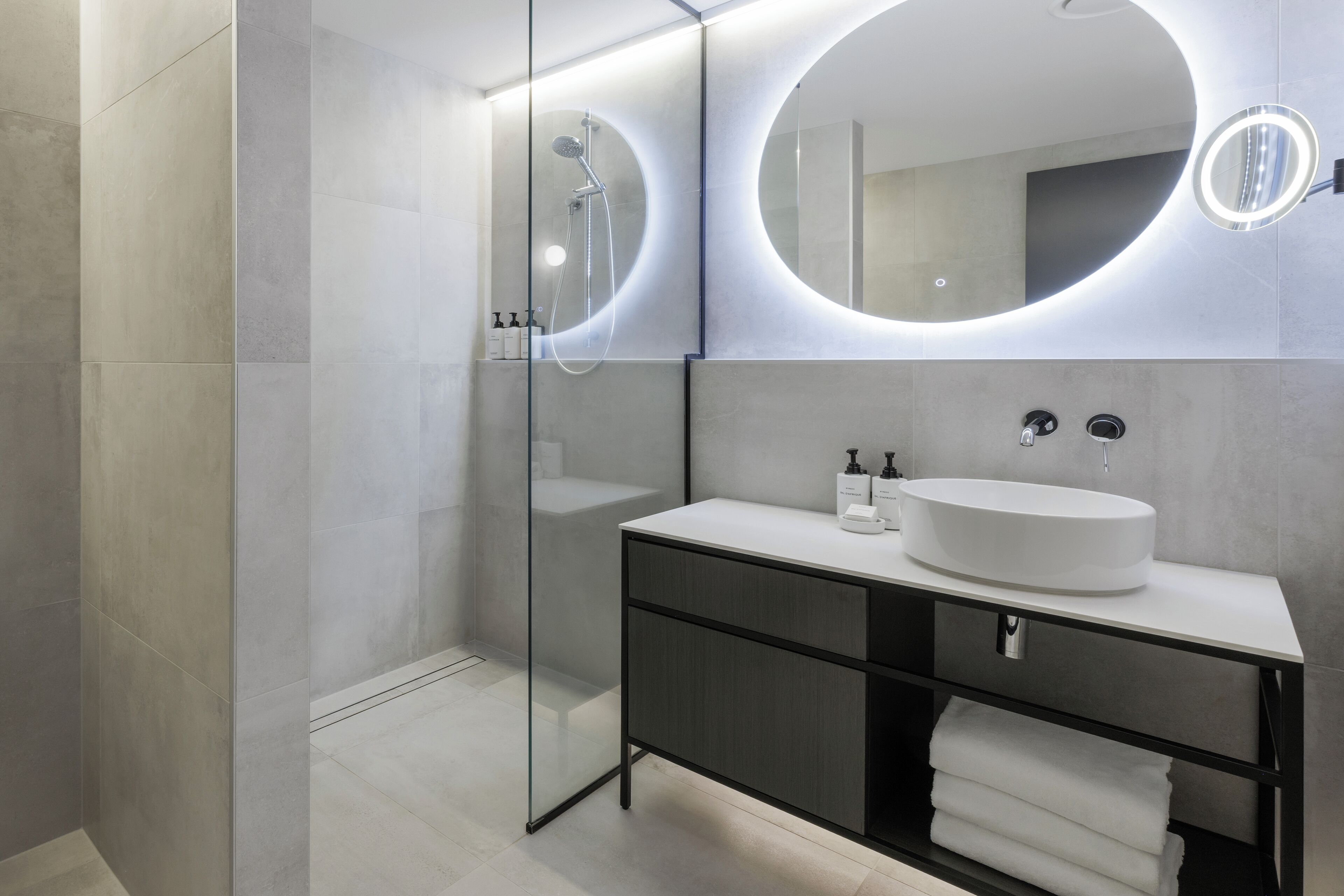 A modern bathroom with a glass shower enclosure, a round mirror with integrated lighting, a white sink, and a dark-toned vanity with a towel rack.