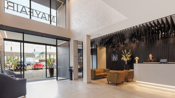 A modern hotel lobby with a reception desk, seating area, and large windows overlooking the street. - The Mayfair (Christchurch)