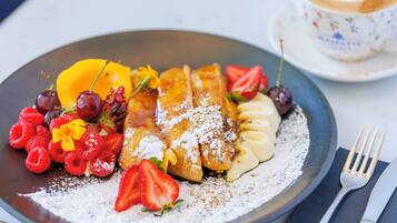 Un plato de comida con diversas frutas y un postre, acompañado de una taza y un tenedor sobre una mesa.