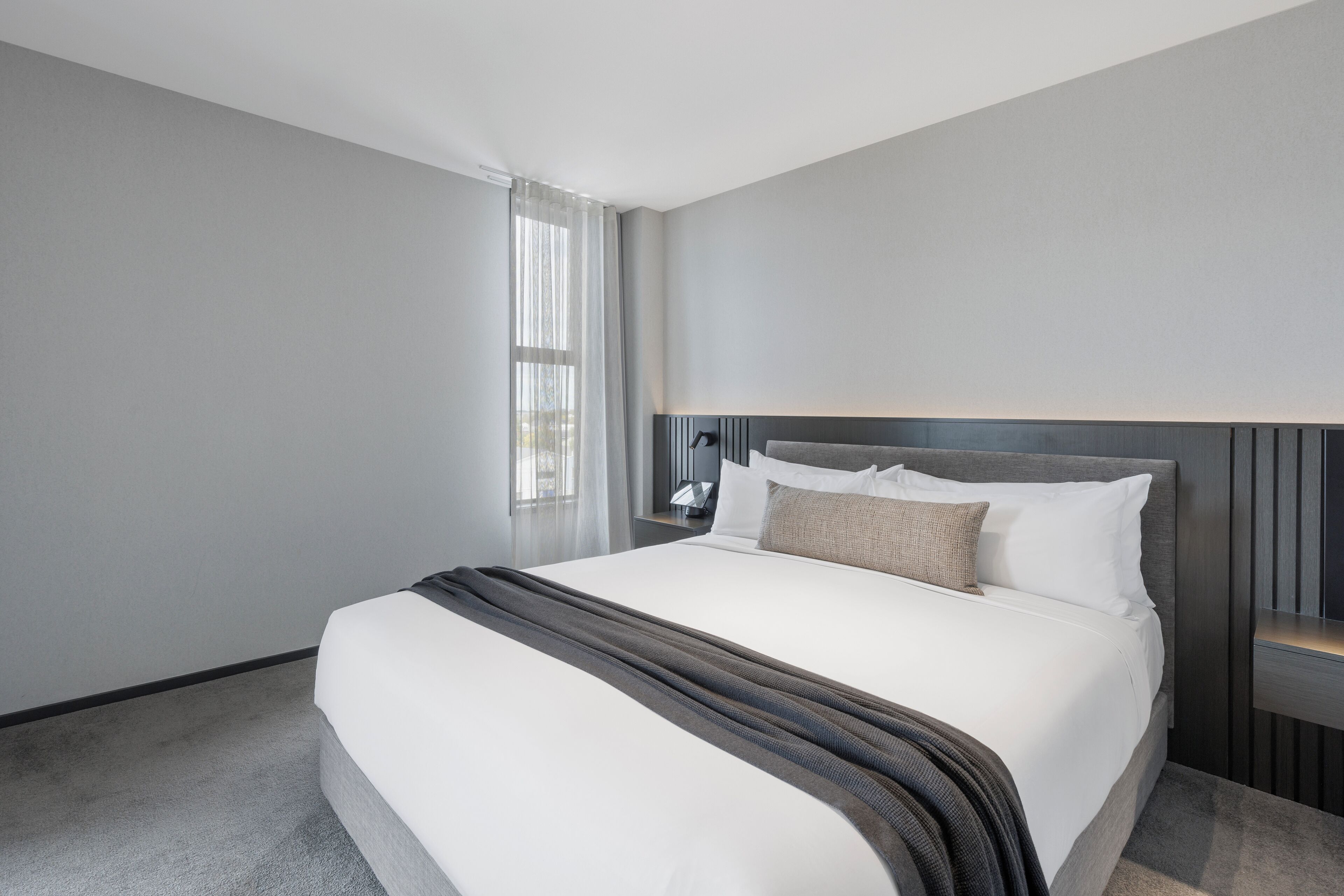 Una habitación de hotel moderna con una cama grande, una mesita de noche y una ventana con cortinas translúcidas.