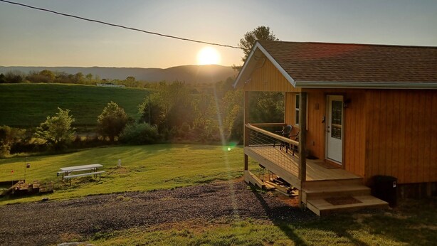 Une cabane en bois avec une véranda, une table de pique-nique et une vue sur le coucher de soleil au-dessus d’un champ verdoyant.