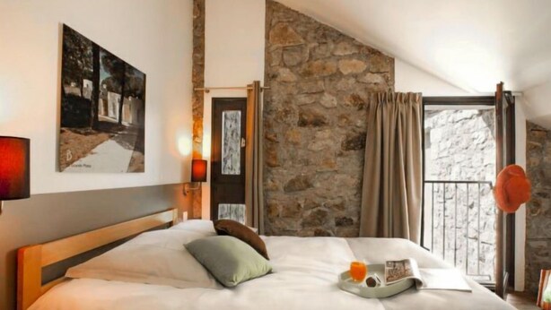 Un dormitorio con pared de piedra, cama con almohadas, mesita de noche con lámpara y ventana con cortinas.