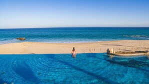 Una persona está de pie en una piscina azul cristalina, con vista al océano y a una playa de arena.