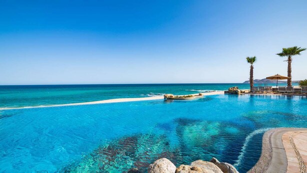 Piscina azul cristalina con vista al océano, rodeada de rocas y palmeras.