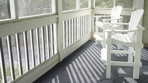 A sunlit porch with white Adirondack chairs and a small table. - Almost oceanfront Pebble Beach condo, indoor & outdoor pools, 3min to the beach! (Emerald Isle)