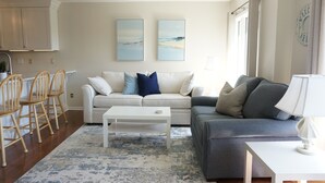 A living room with a white sofa, a grey sofa, a coffee table, and wooden chairs. There are two paintings on the wall and a window with curtains. - Almost oceanfront Pebble Beach condo, indoor & outdoor pools, 3min to the beach! (Emerald Isle)