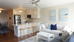A modern kitchen with white cabinets, a stainless steel refrigerator, and a microwave. There is a dining area with a table and chairs. The living room has a gray sofa and a white coffee table. - Almost oceanfront Pebble Beach condo, indoor & outdoor pools, 3min to the beach! (Emerald Isle)