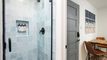 A modern bathroom with a glass shower enclosure, a sink with a soap dispenser, and a door leading to a dining area with a table and chairs.