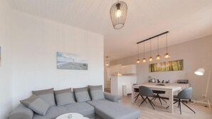 Un salon moderne avec un canapé d’angle gris, une petite table blanche et une cuisine en arrière-plan.