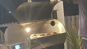 Una griglia per barbecue con display digitale e pannello di controllo, posizionata all'aperto con una recinzione in legno e una pianta in vaso sullo sfondo.