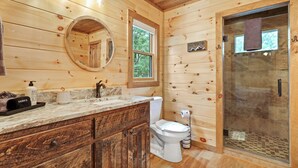 Baño de cabaña de madera con tocador de madera, inodoro, cerramiento de ducha de vidrio y ventana.