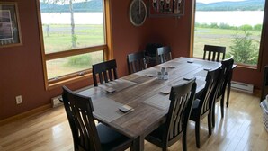 A dining room with a large wooden table, six chairs, and a view of a lake through large windows. - Beautiful Lake House for Rent (Neebing)