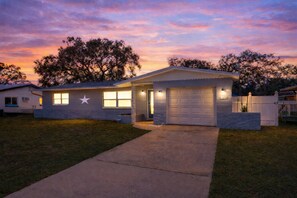 A single-story house with a front garage, a star on the wall, and a well-lit driveway at dusk. - Beach Escape: King Bed | Games | Sunroom | Bikes (Largo)