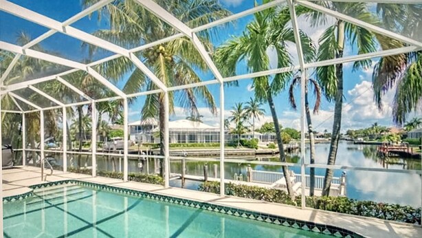 A pool area with a glass enclosure, palm trees, and a water body. - 8 Minute Walk to Beach Luxury 4 Bdrm Waterfront home -salt water heated Pool (Marco Island)