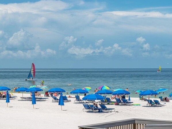 Relax on the beach with shade from colorful umbrellas, a sailboat on the calm sea, and a lifeguard stand nearby.