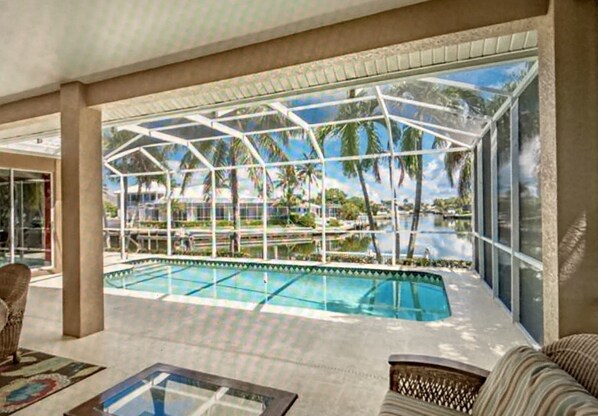 A pool area with a glass enclosure, lounge chairs, and a view of palm trees and a pool.