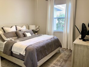 A neatly made bed with a grey and white color scheme, a bedside table with a lamp, and a window with blinds.