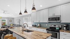 A modern kitchen with white cabinets, a granite countertop, a stainless steel sink, a microwave, an oven, and a coffee maker. There are two bar stools at the island. - Luxurious Private Pool | Beach Walk with Private Access 🏖️✨ (Miramar Beach)