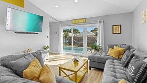 A modern living room with grey sofas, a flat-screen TV, a 'WELCOME' sign, and a pool view. - Luxurious Private Pool | Beach Walk with Private Access 🏖️✨ (Miramar Beach)