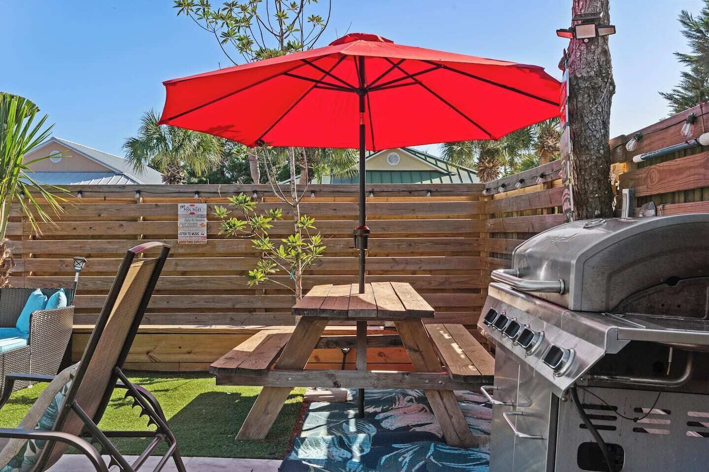 A backyard with a wooden table and bench, a red umbrella, and a barbecue grill.