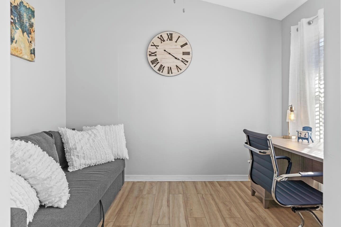 A modern room with a grey sofa, a wooden floor, a desk with a chair, and a wall clock.