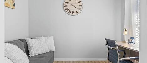 A modern room with a grey sofa, a wooden floor, a desk with a chair, and a wall clock.