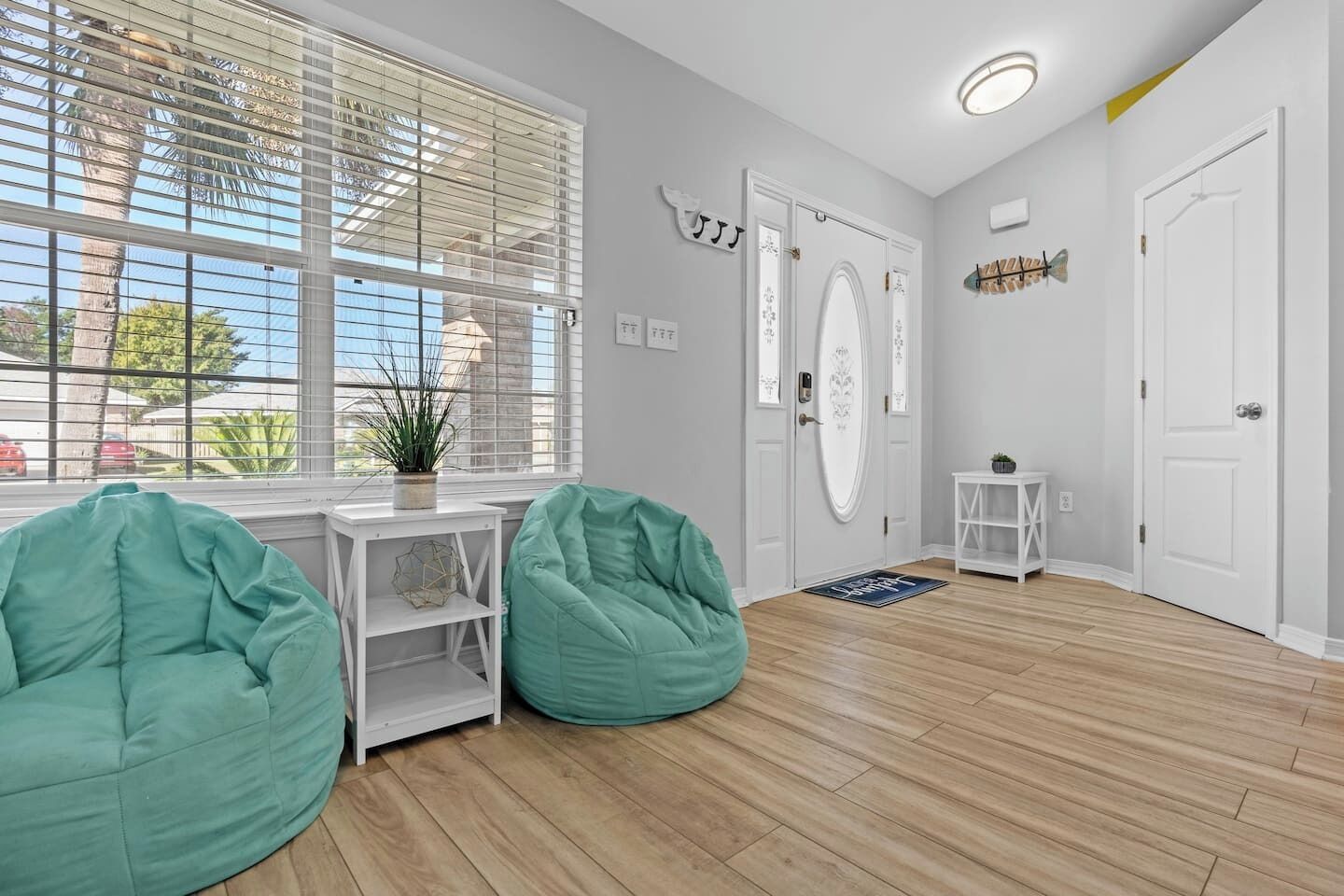 A room with a wooden floor, a white door, a window with blinds, and a small white table with a plant.