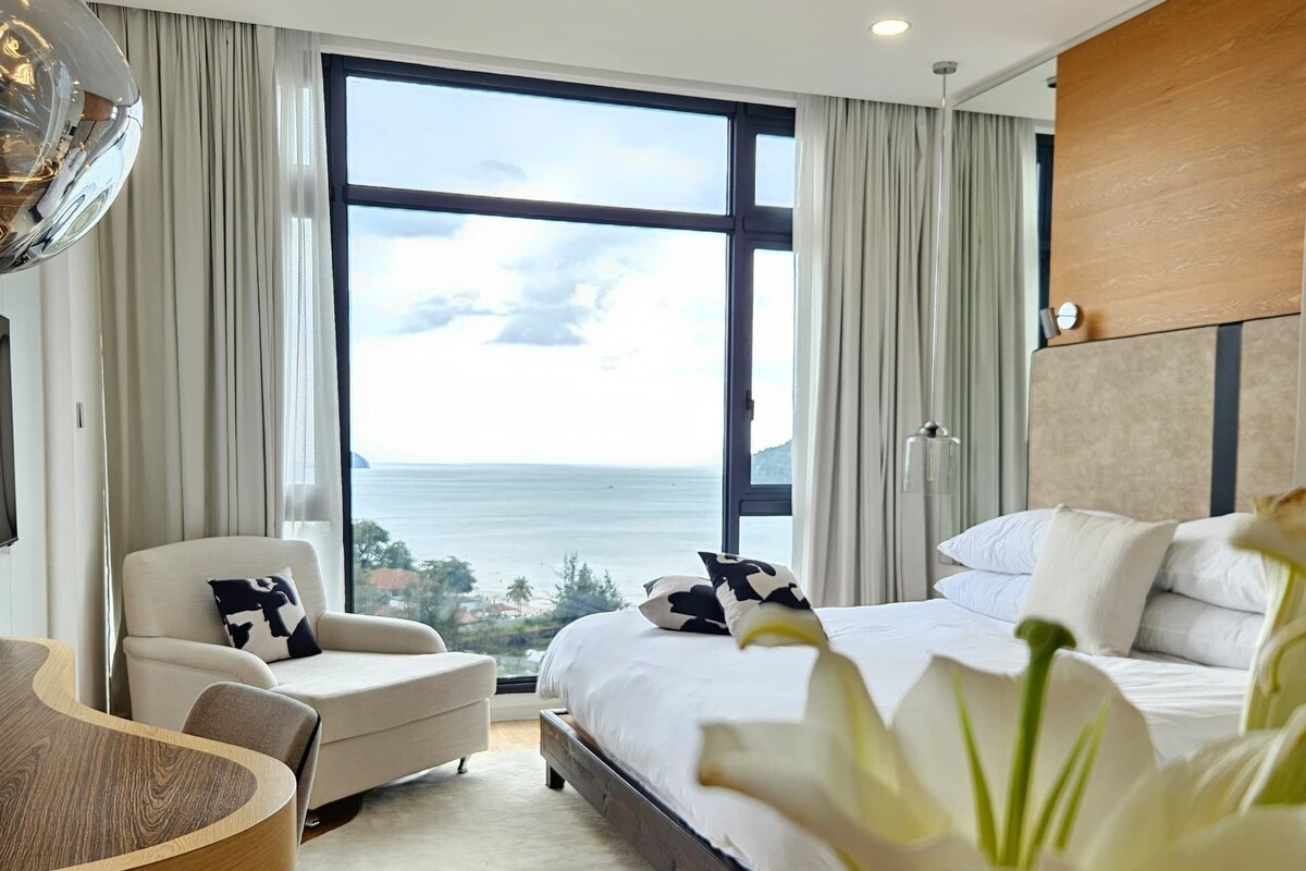 A modern hotel room with a large bed, a seating area with a table, and a view of the sea.