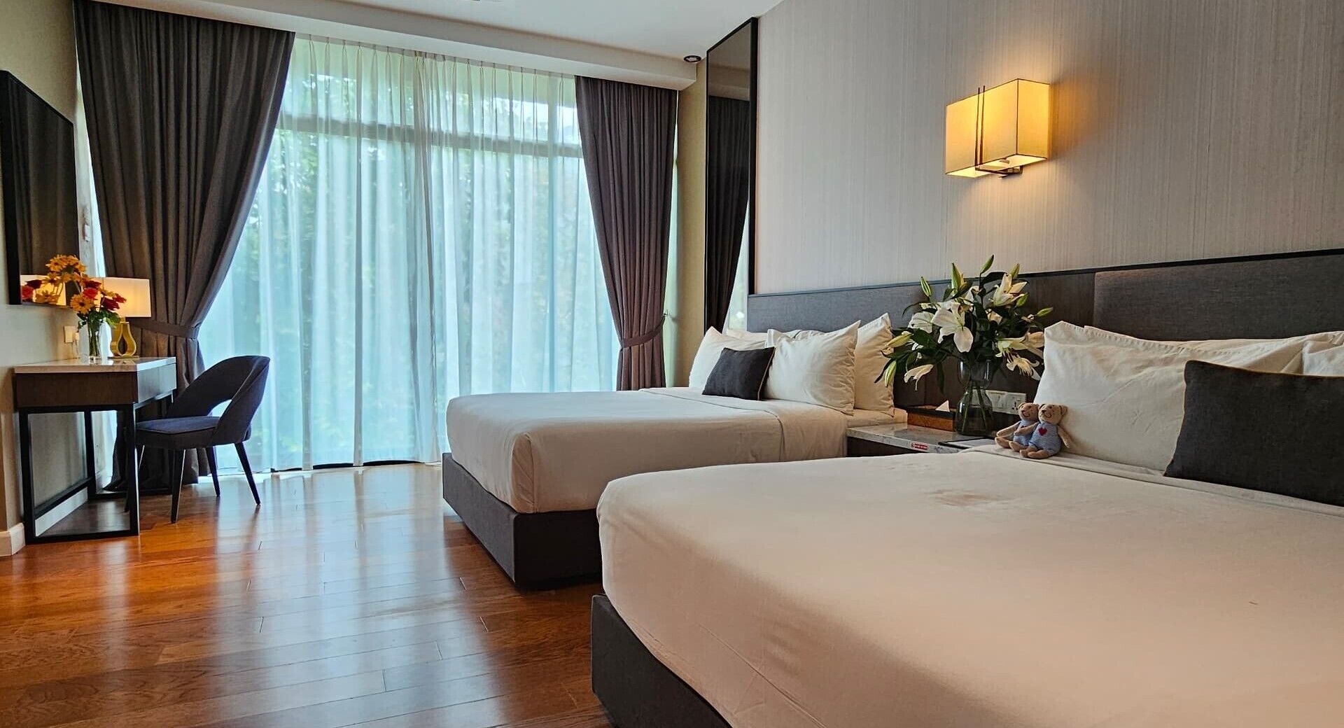 Una camera d'albergo con due letti, una scrivania, una sedia e un'ampia finestra con tende.