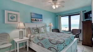 Ein Schlafzimmer mit einem Bett, zwei Nachttischen, einem Sessel, einem Deckenventilator, einem Fernseher und Blick auf den Strand.