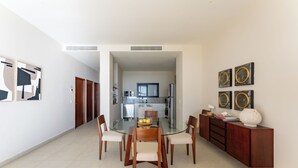 A modern dining area with a glass table, wooden chairs, and a kitchen in the background. - Medusa🌴 Elegant 1BDR Apartment by the Sea | Luxury & Relaxation in Tulum (TULUM)